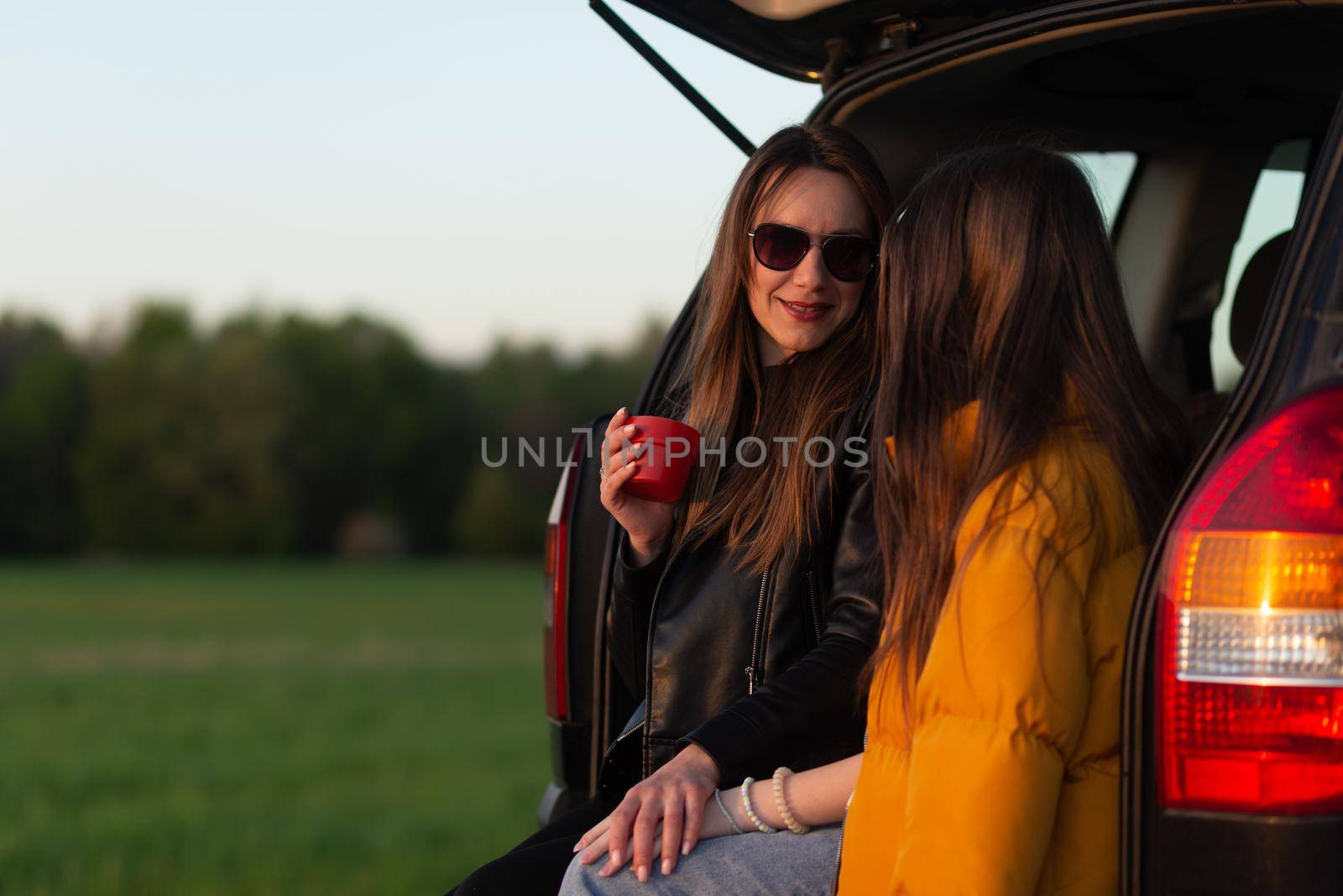 Mother and daughter camping on a hill and admiring the sunset while sitting in the car trunk
