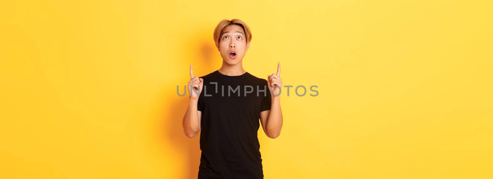 Portrait of curious and amazed asian guy with blond hair, wearing black t-shirt, looking and pointing fingers up astonished, yellow background.