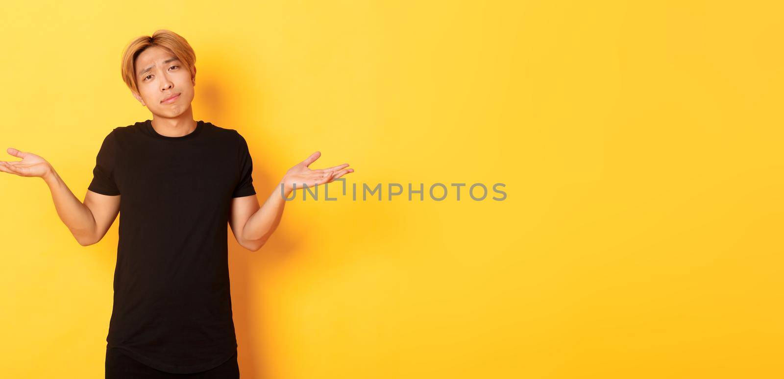 Portrait of clueless asian blond guy, wearing black clothes, shrugging and looking puzzled, standing yellow background by Benzoix