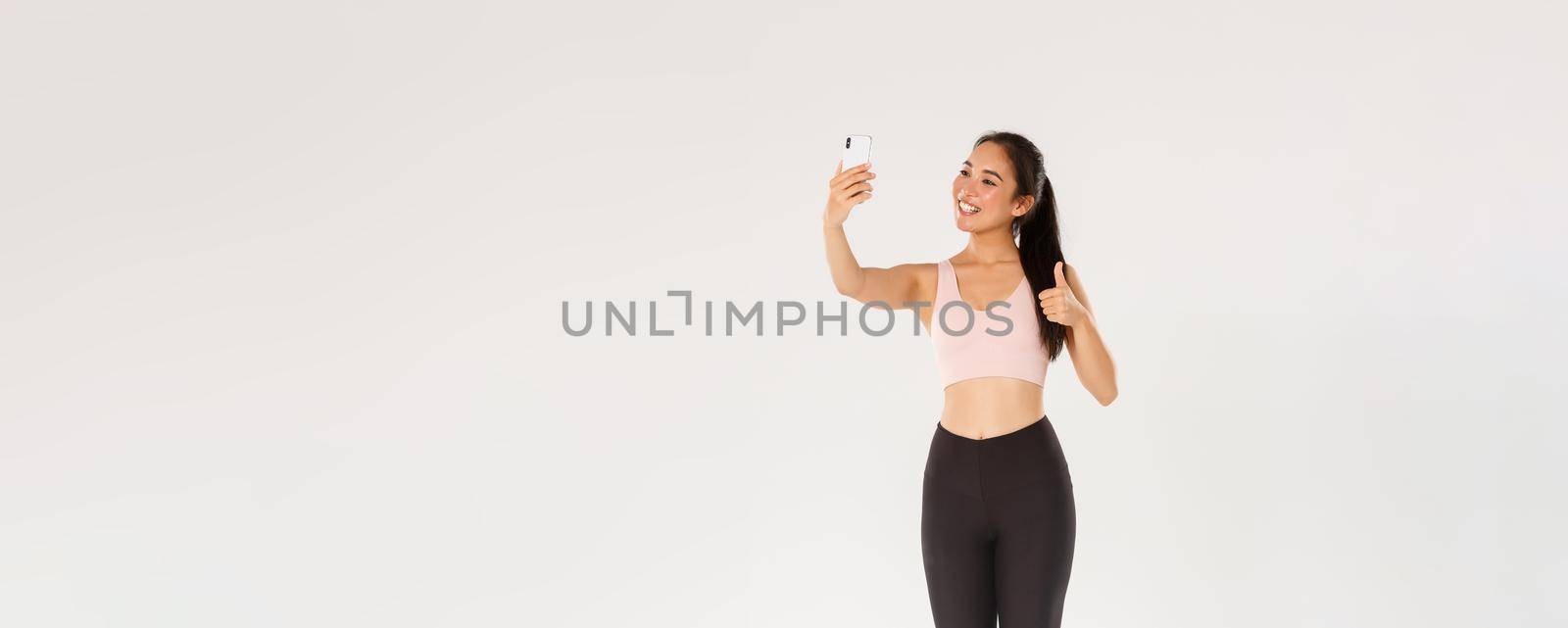 Full length of smiling female lifestyle blogger, asian fitness girl in sportswear taking selfie in gym, showing thumbs-up, demonstrate workout exercies for followers online, live stream from training by Benzoix