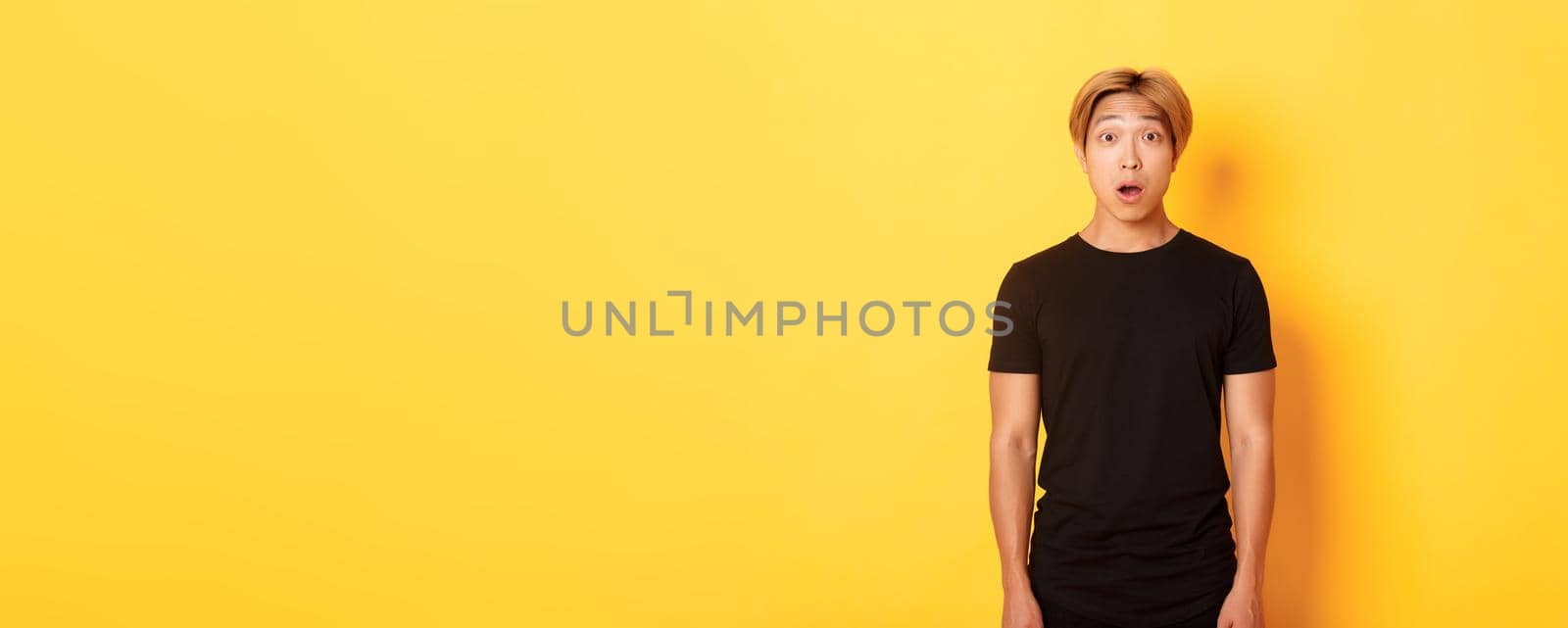 Portrait of startled handsome asian blond guy, drop jaw and gasping impressed, yellow background.