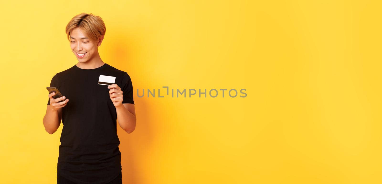 Smiling handsome asian man paying for online purchase, shopping in internet, holding smartphone and credit card, yellow background.