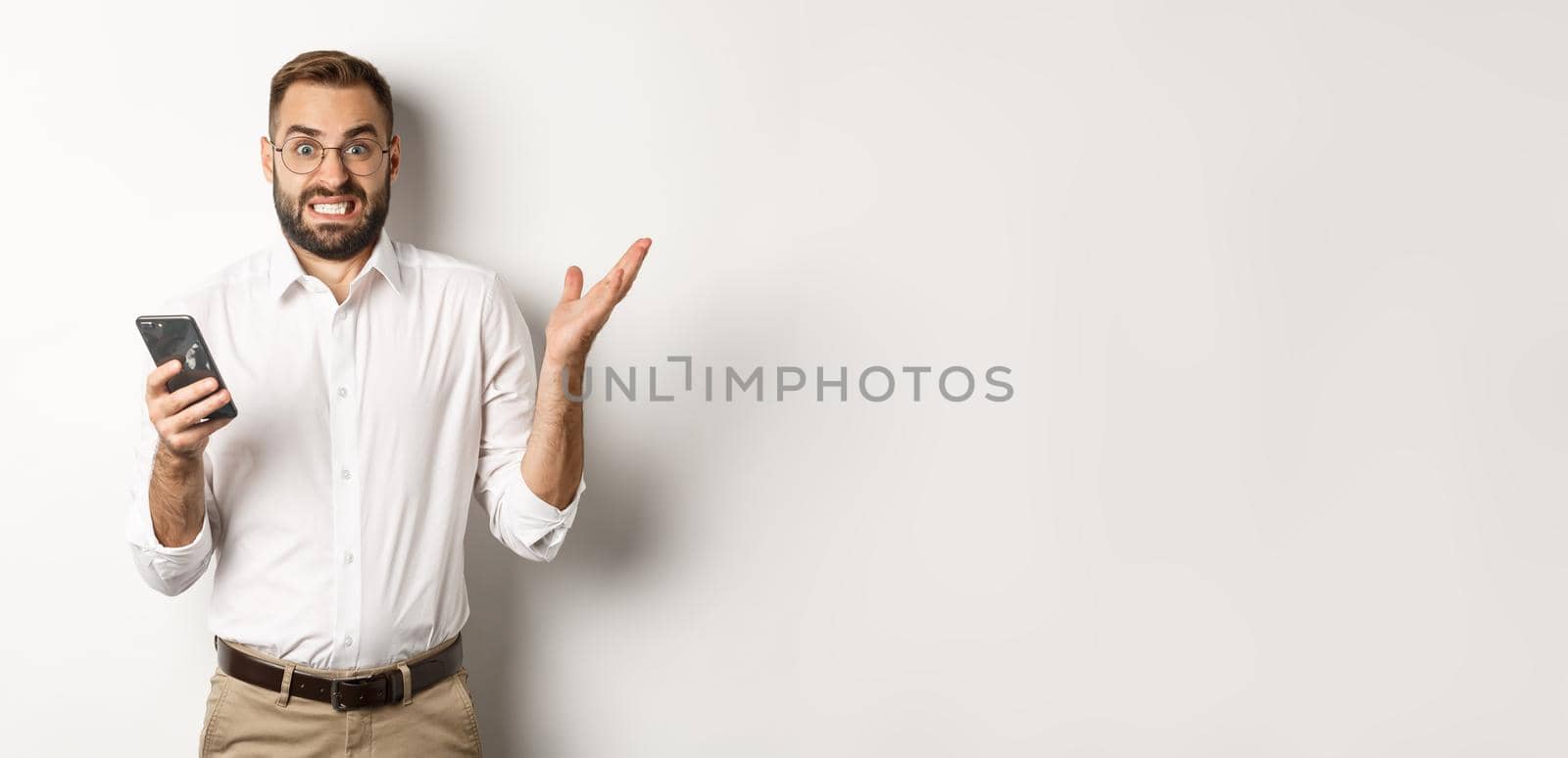 Confused businessman reading strange message on mobile phone, looking annoyed, standing over white background.