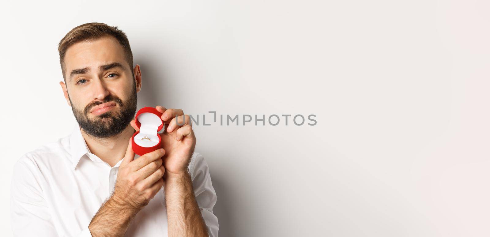 Close-up of hopeful man begging to marry him, looking sad and showing wedding ring, making a proposal, white background.