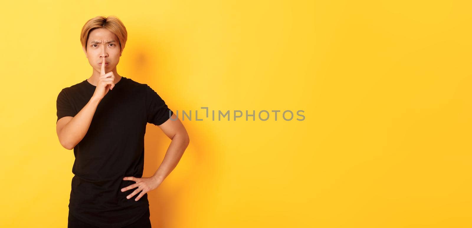 Portrait of angry asian man frowning, shushing at camera disappointed, standing yellow background.