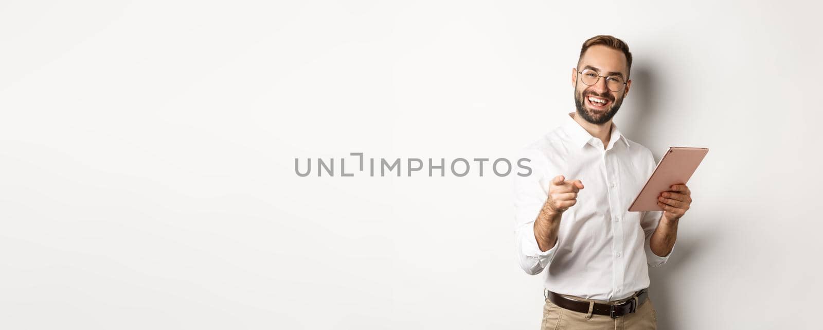 Happy and satisfied boss praising good job, reading on digital tablet and pointing at you camera, standing over white background by Benzoix