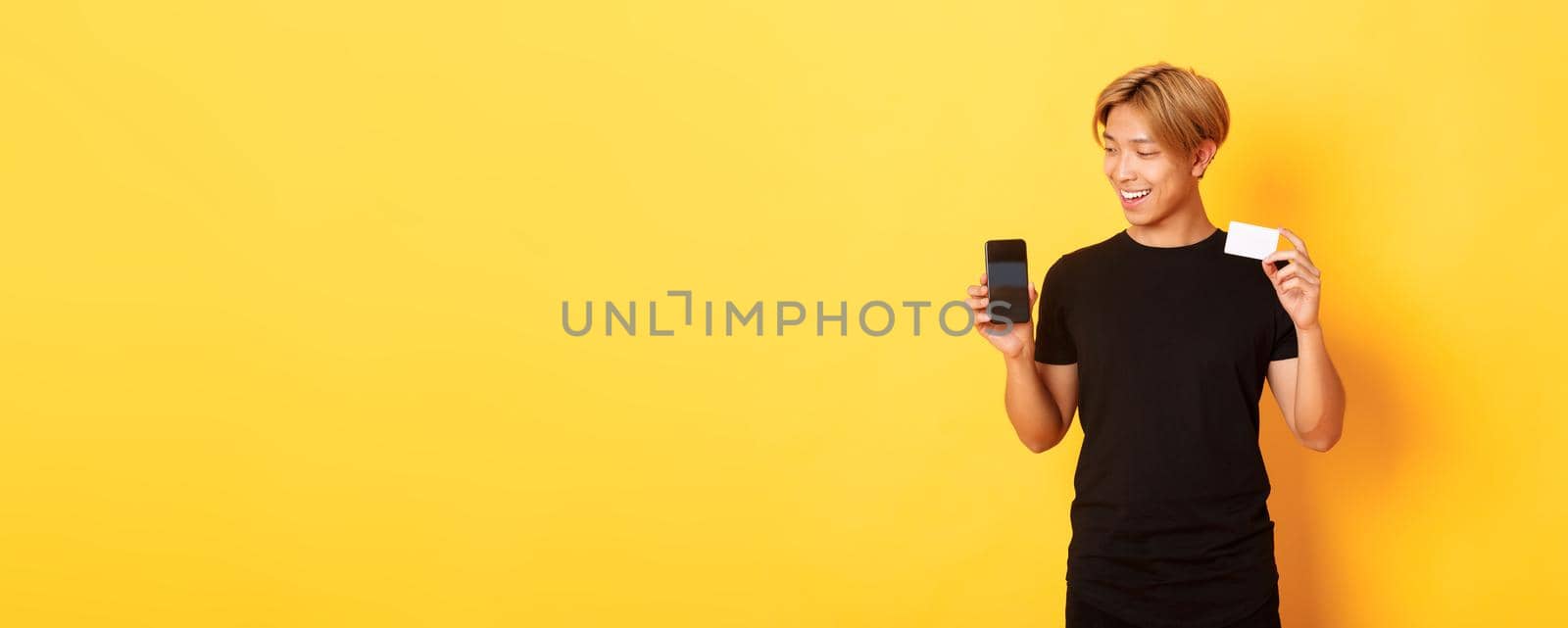 Portrait of pleased handsome asian guy, showing smartphone screen and credit card, looking at mobile phone display, yellow background by Benzoix