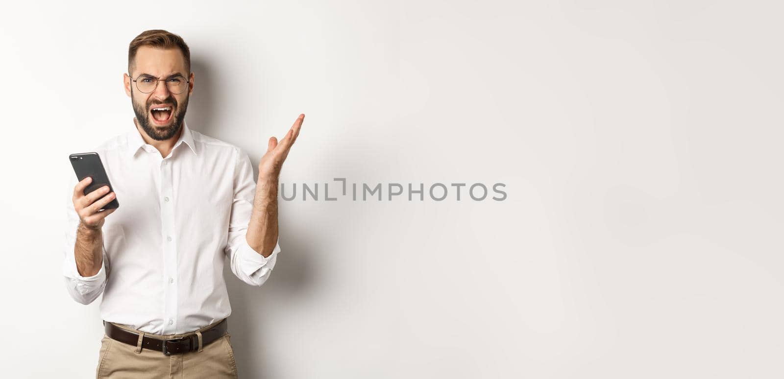 Frusteated man using mobile phone and looking disappointed, complaining, standing over white background.