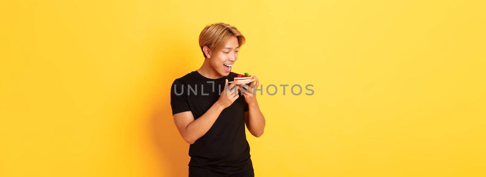 Joyful handsome asian guy smiling and looking tempted at cake while making bite, standing yellow background by Benzoix