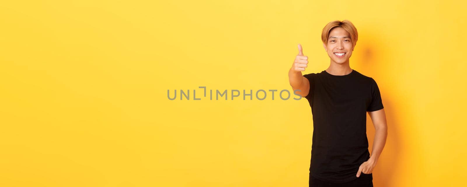 Portrait of satisfied asian man with blond hair, standing over yellow background and showing thumbs-up.