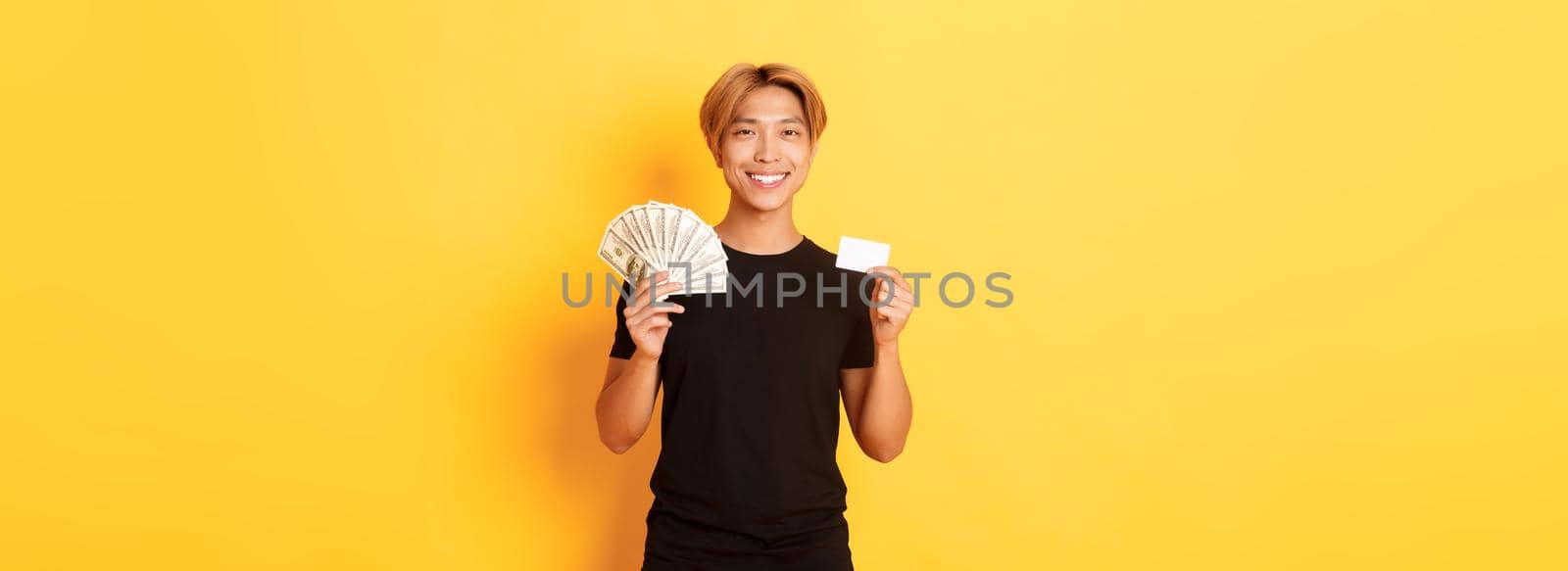Pleased handsome asian guy showing money and credit card, smiling happy, standing yellow background by Benzoix