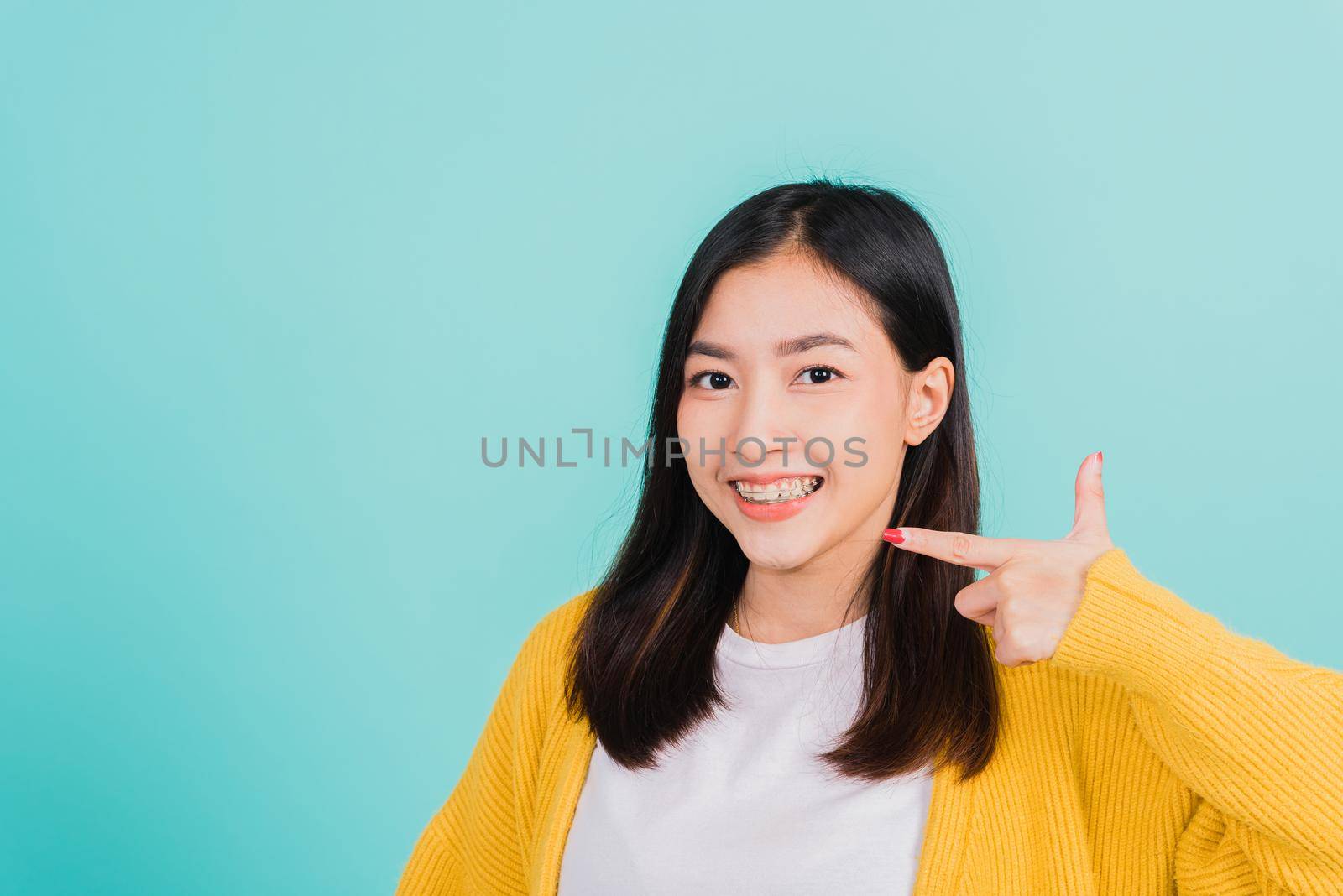 Asian beautiful young woman teen pointing finger to teeth, portrait of happy Thai female confident smiling show white teeth, studio shot isolated on blue background, Dental health care concept