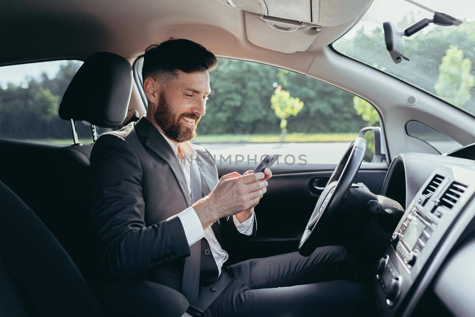 Young man with a beard businessman driving a car in the parking lot smiles and writes on the phone reads the news successful man