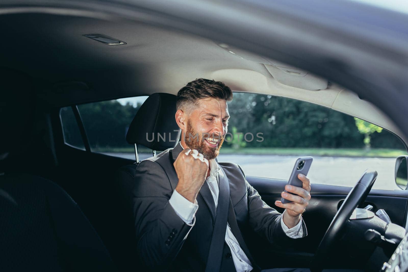 Male businessman successful rejoices and smiles reads news from mobile phone win and success meeting, man sits in car