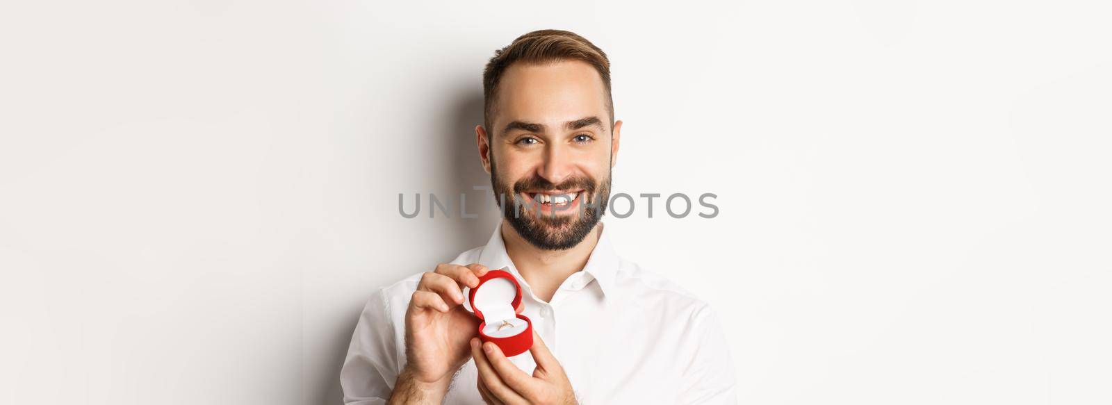 Close-up of happy handsome man making a proposal, holding wedding ring in box and smiling, asking to marry him, white background.