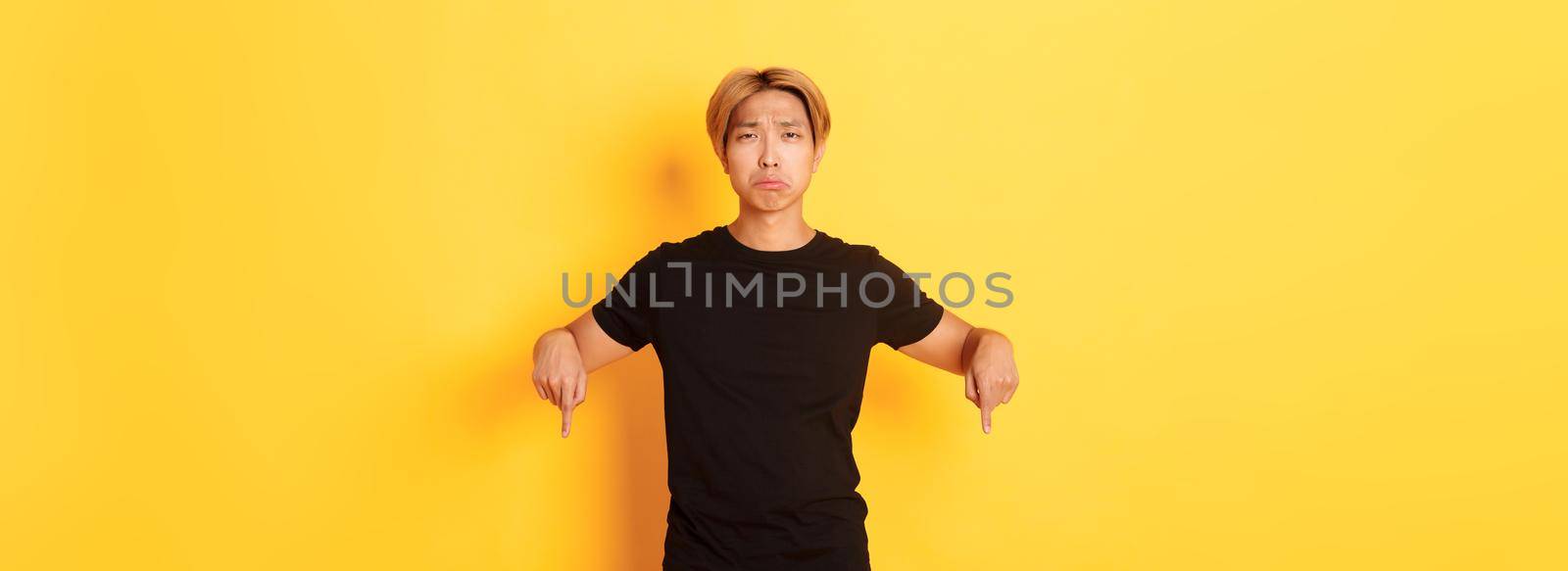 Portrait of disappointed gloomy asian guy, pouting upset and pointing fingers down with regret and uneasy feeling, yellow background by Benzoix