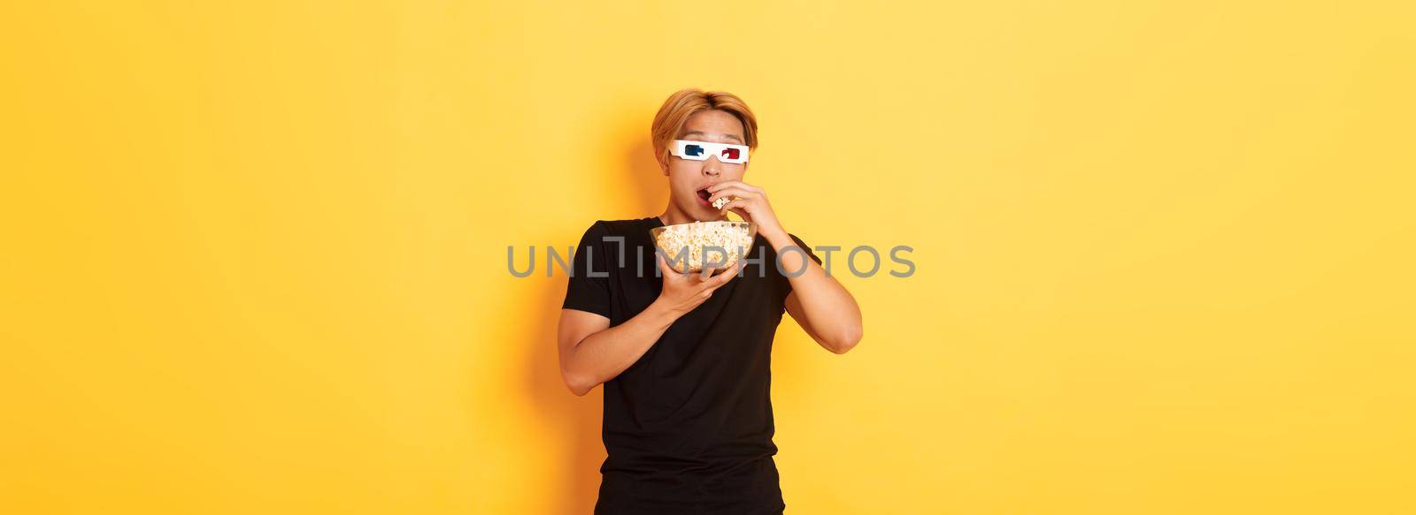 Impressed and thrilled asian guy attend cinema, watching movie or tv series in 3d glasses, eating popcorn and looking shocked, standing yellow background.