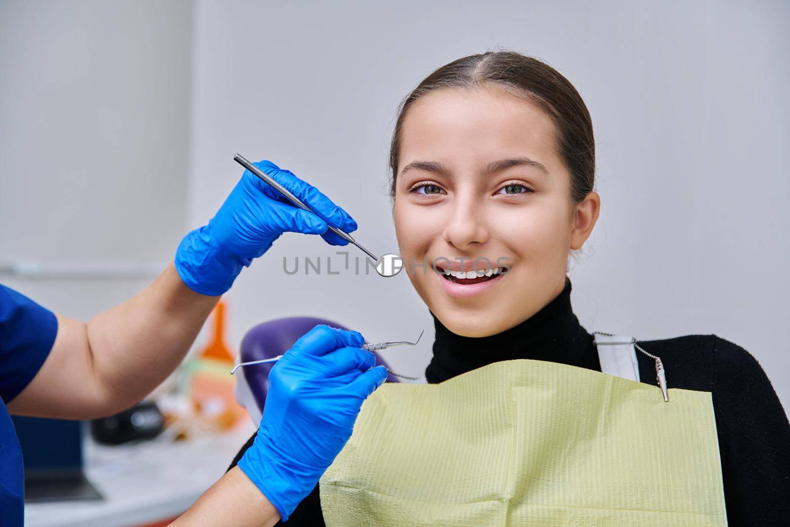 Portrait of young teenage girl in dental chair with hands of doctor with tools. Female teenager smiling with teeth looking at camera in dentist office. Adolescence hygiene treatment dental health care