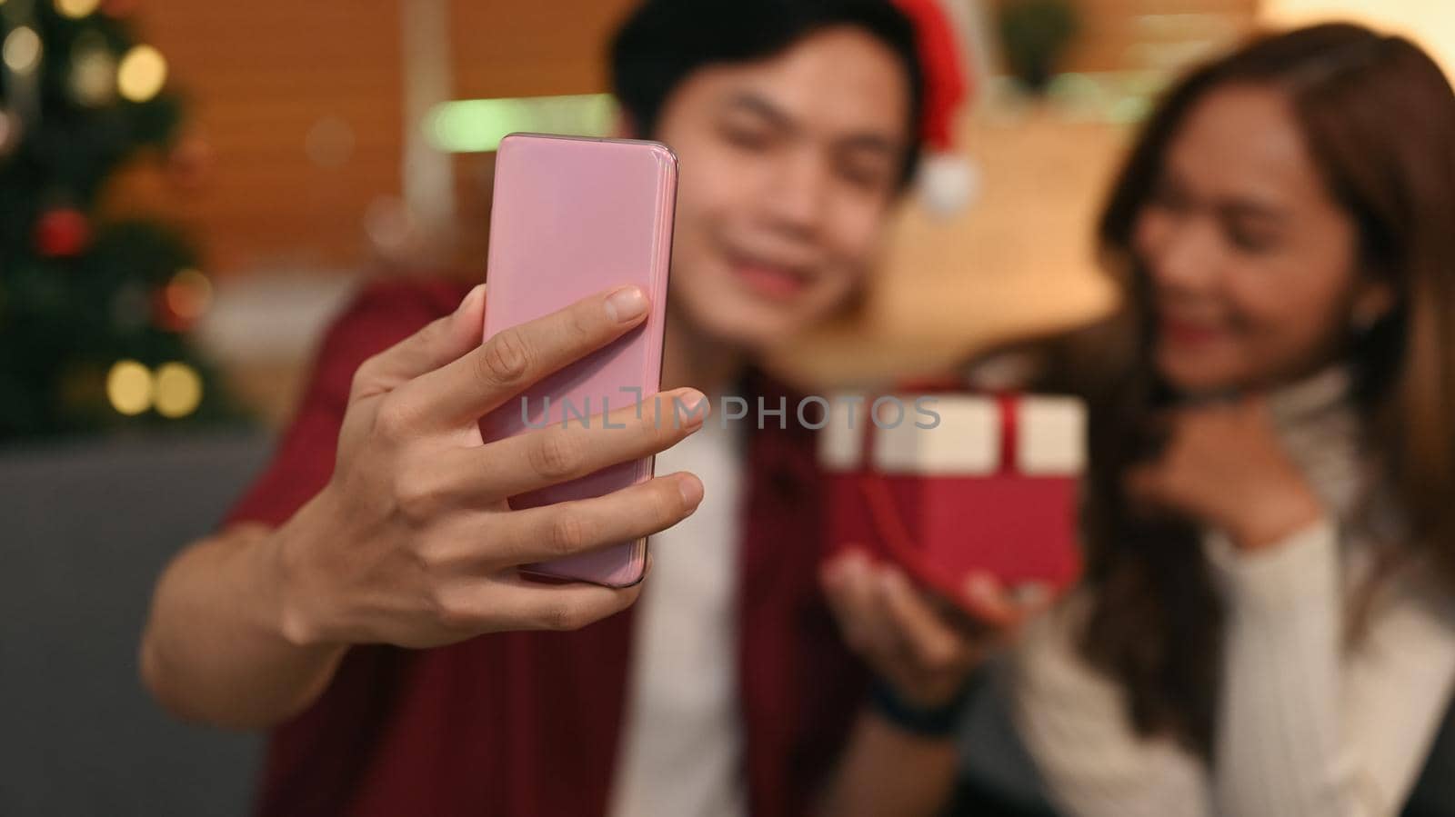 Happy young couple in Santa hat taking selfie on mobile phone while sitting in decorated room. Celebration, holidays and people concept by prathanchorruangsak
