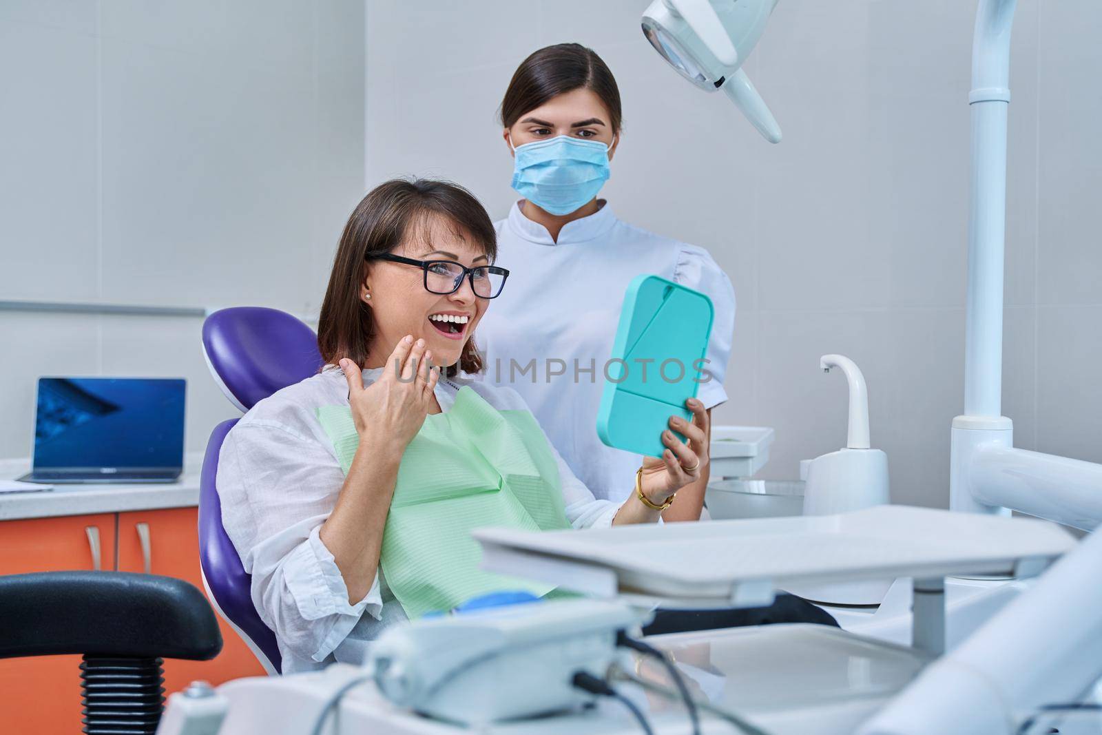 Happy middle aged woman together with dentist, patient sitting in dental chair looking at mirror. Prosthetics, treatment, implantation, dental teeth health, beauty care concept
