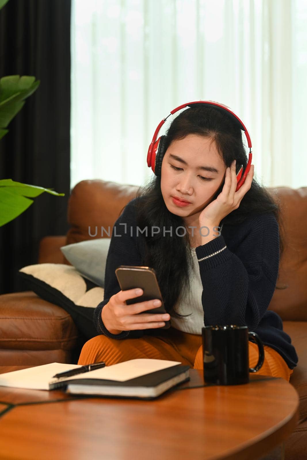 Pretty young asian woman wearing wireless headphone and using mobile phone on couch at home by prathanchorruangsak