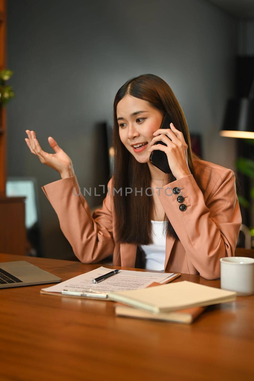 Confident saleswoman talking on mobile phone with her client, sitting at contemporary office by prathanchorruangsak