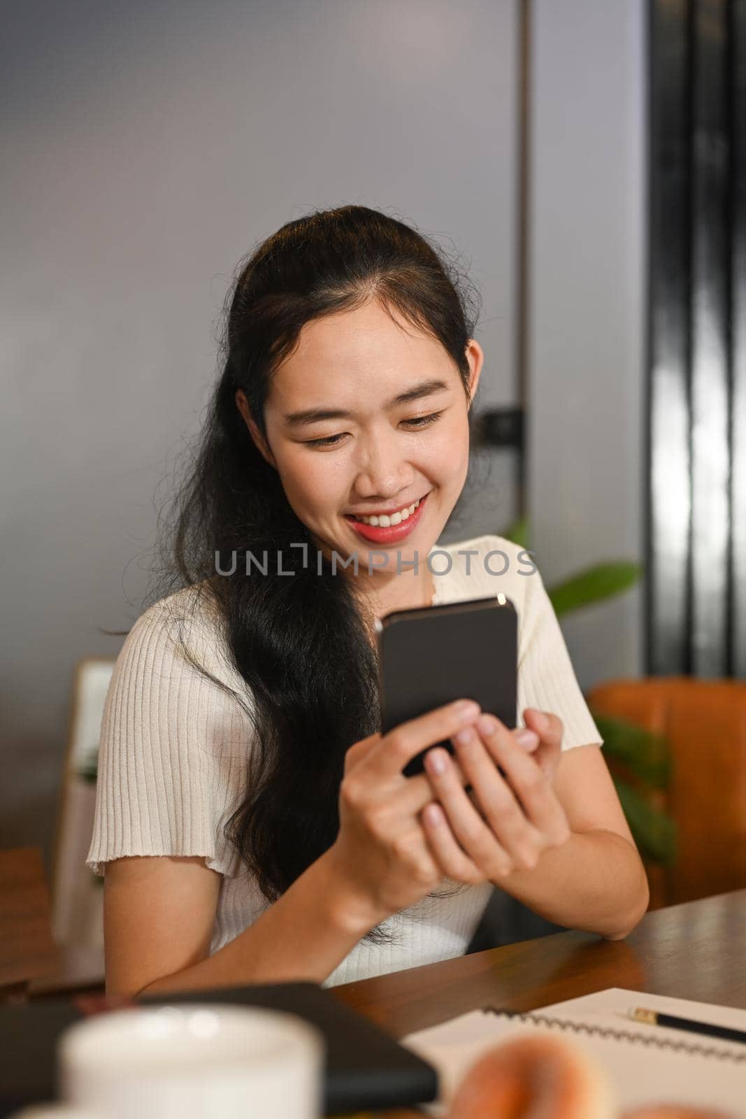 Smiling young woman sitting in her working desk and reading text on her mobile phone.