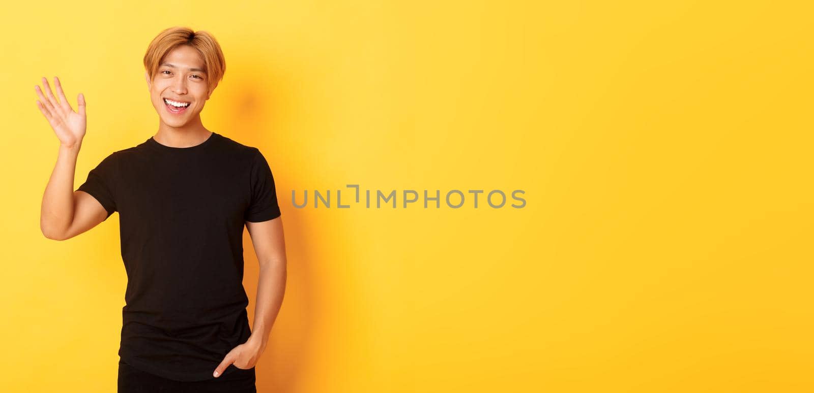 Portrait of handsome friendly asian guy in black outfit, waving hand to say hello and smiling, greeting someone, standing yellow background.