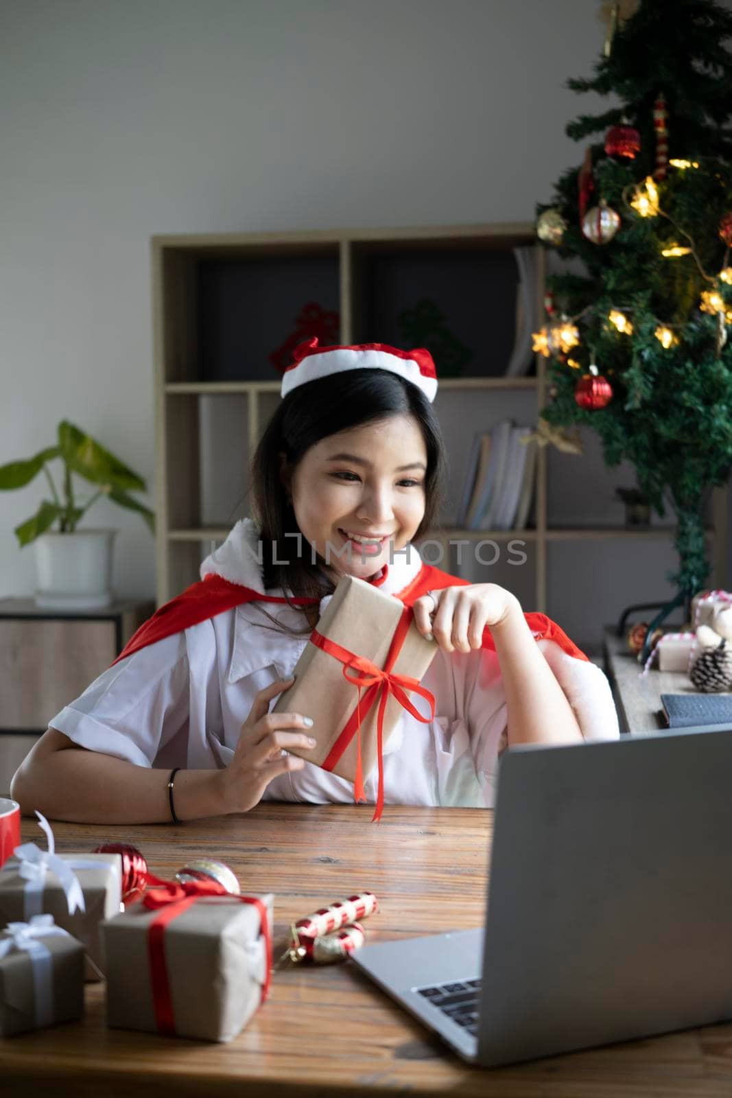 Cheerful asian woman holding gift box and having video call via laptop computer at home. by prathanchorruangsak