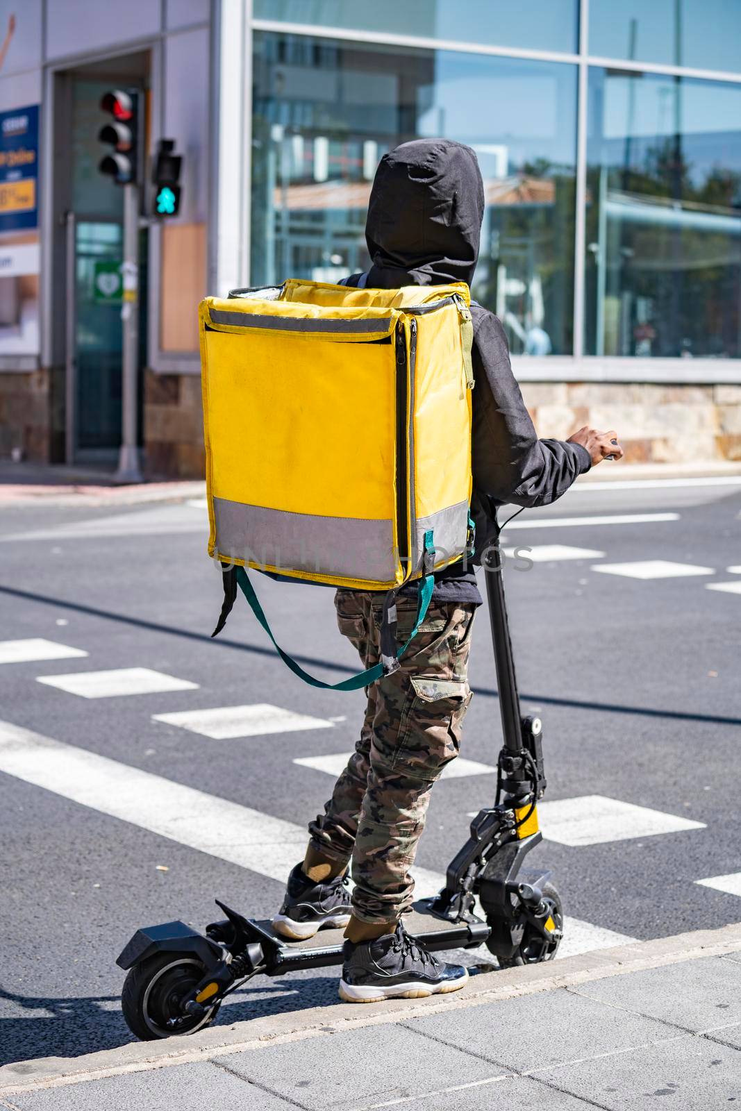 Man courier online food delivery with yellow thermal backpack waiting the green traffic light on an electric scooter. by papatonic