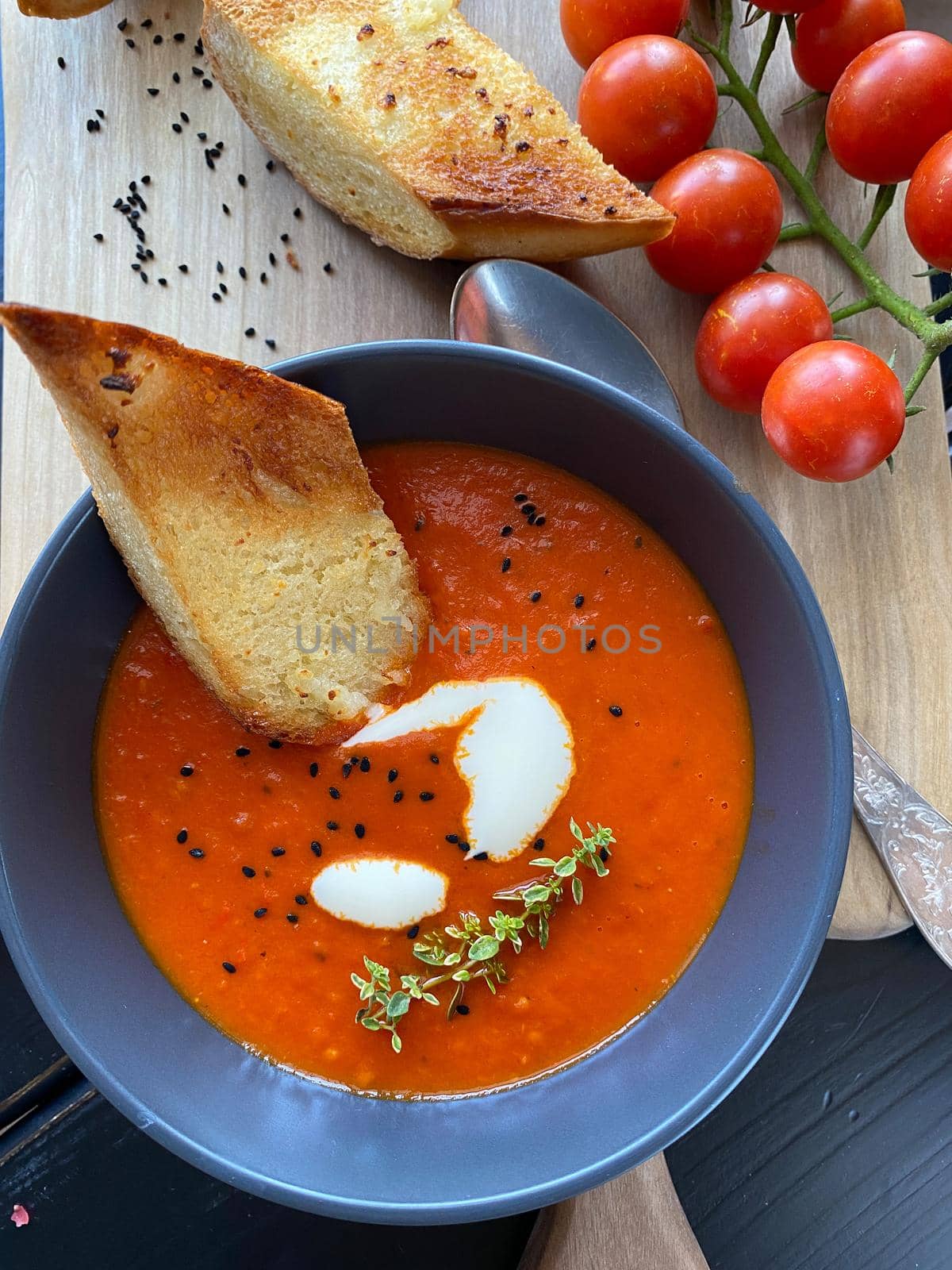 Tomato soup with thyme in a black bowl. Black background and wooden cutting board. Close-up. Top view . Pieces of delicious garlic bread, a branch of tomatoes and a silver spoon. Close up. by Proxima13