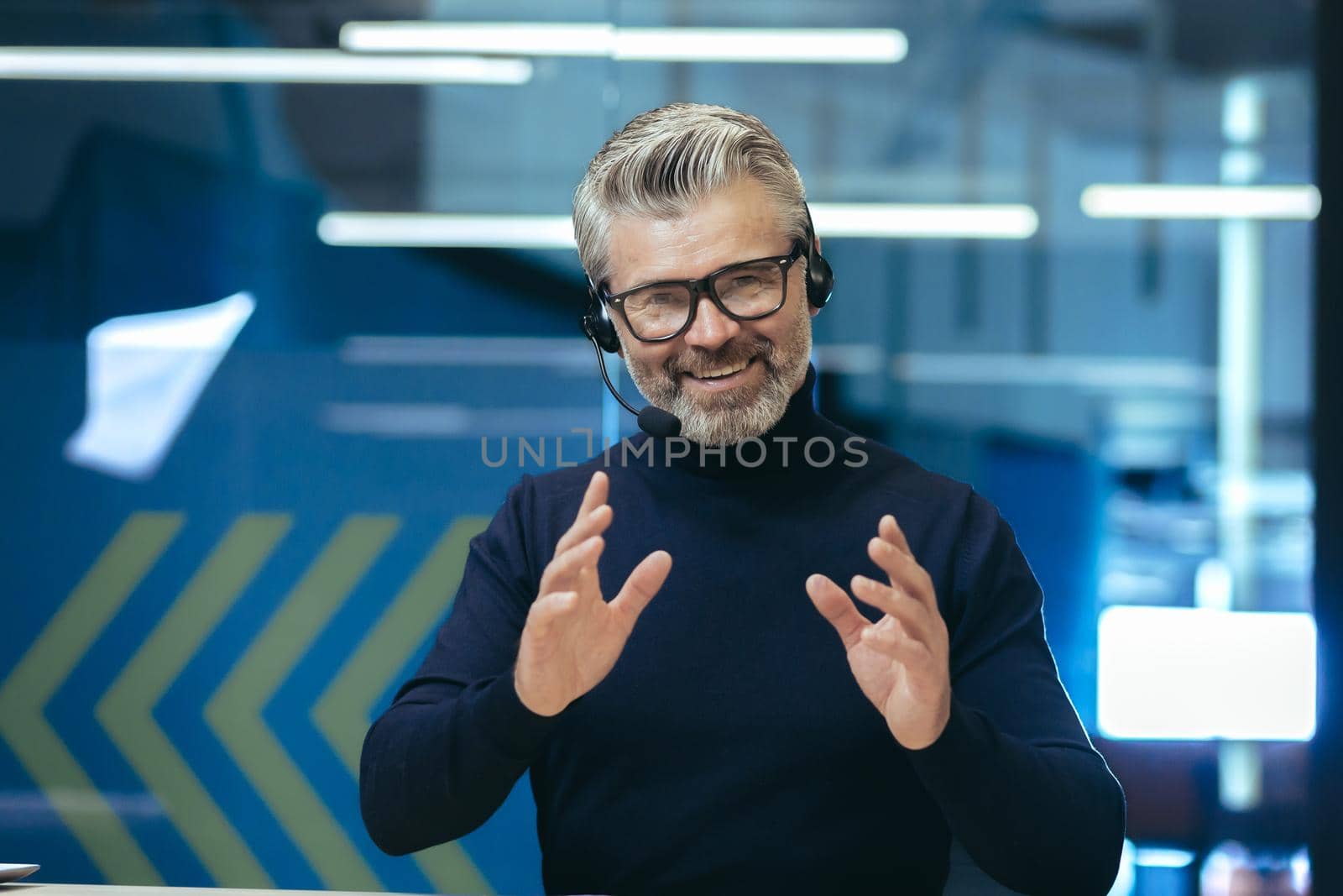 Senior gray haired businessman talking on video call, using headset investor in glasses smiling and looking at camera, webcam view man working inside modern office building.