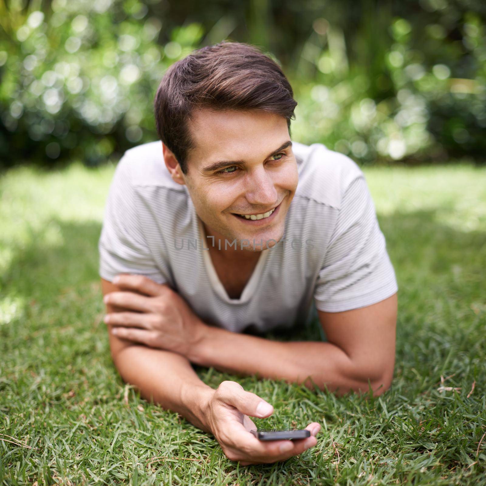 Thinking of the perfect response. a handsome young man using a cellphone while lying outdoors. by YuriArcurs