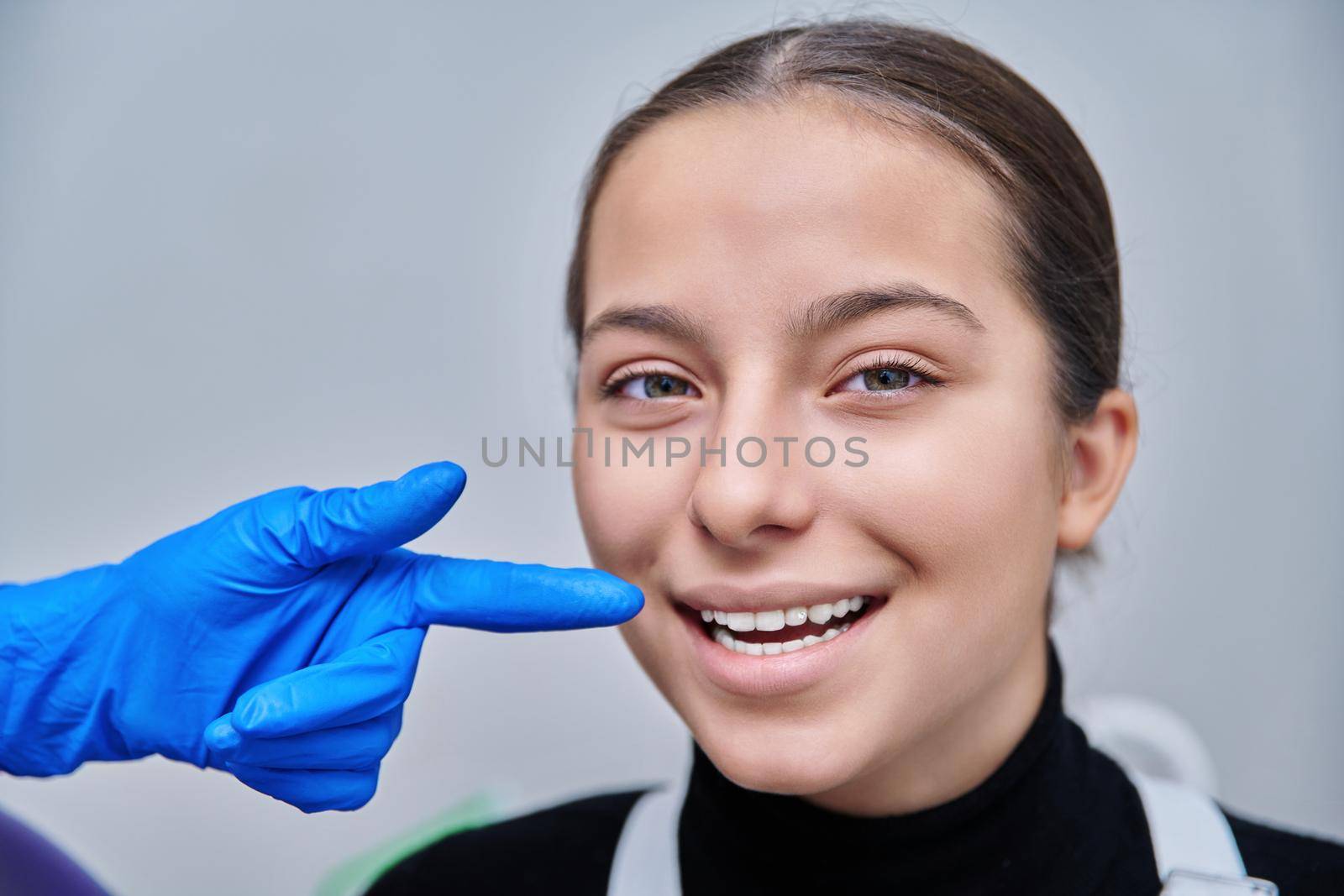 Close-up face smiling young teenage female, dentist hands showing healthy beautiful teeth with luxury jewelry rhinestone dental diamond on patient tooth. Beauty, dentistry, fashion concept