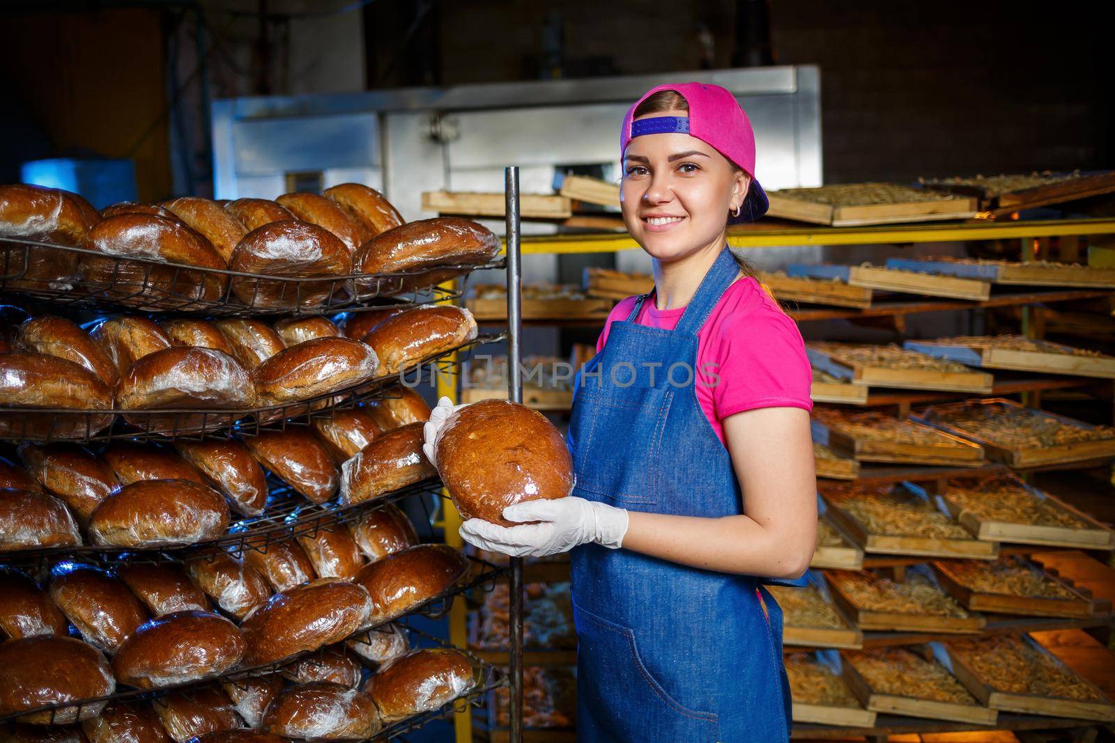 Smiling baker girl in apron holding bread and looking at camera. Young woman is a chef holding fresh baking. A happy girl of European appearance is smiling at the bakery.