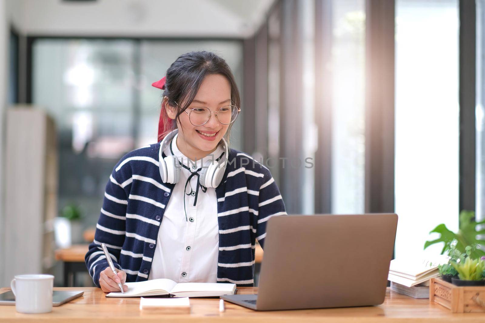 Young adult happy smiling Hispanic Asian student wearing headphones talking on online chat meeting using laptop in university campus or at virtual office. College female student learning remotely..