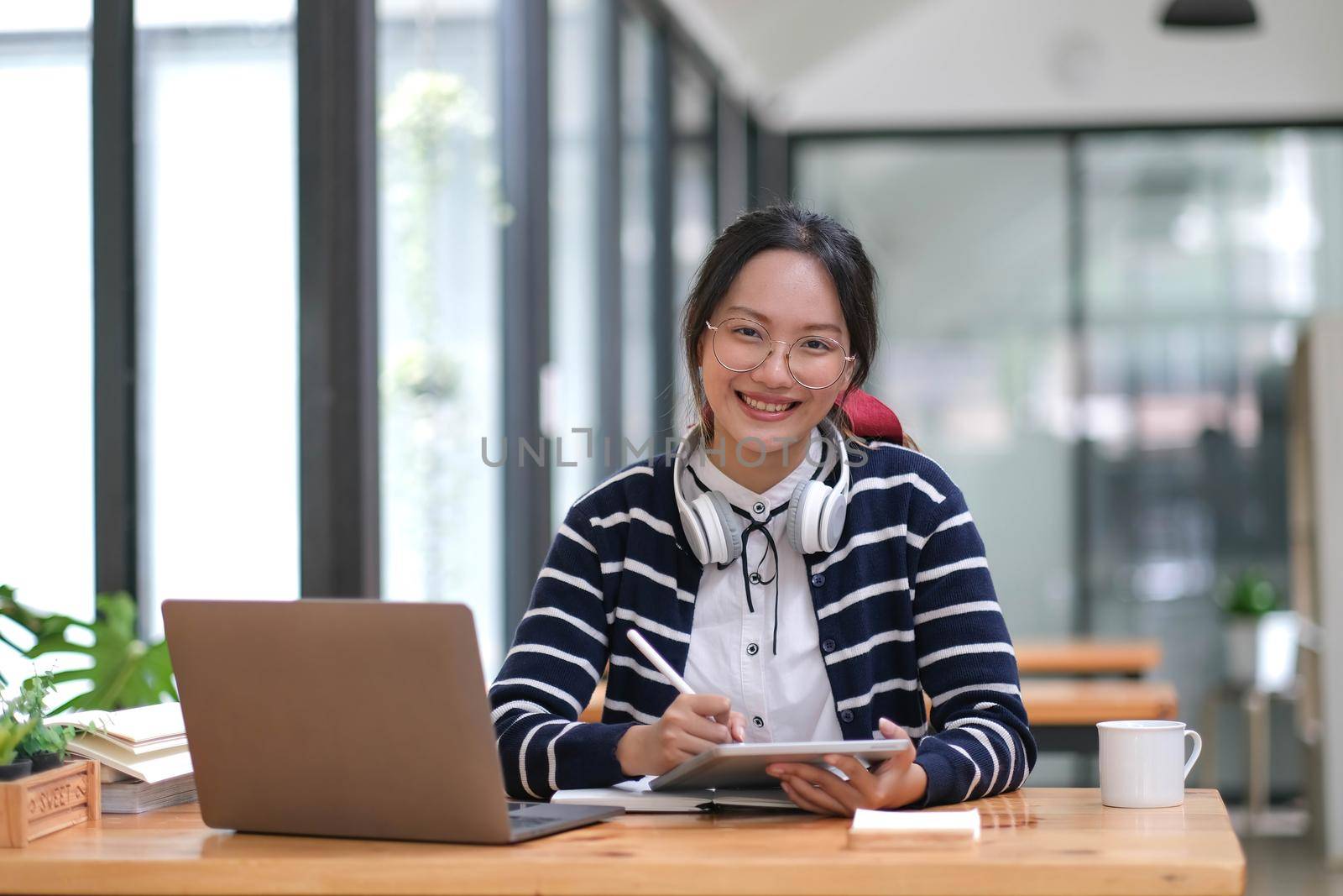 Young adult happy smiling Hispanic Asian student wearing headphones talking on online chat meeting using laptop in university campus or at virtual office. College female student learning remotely..