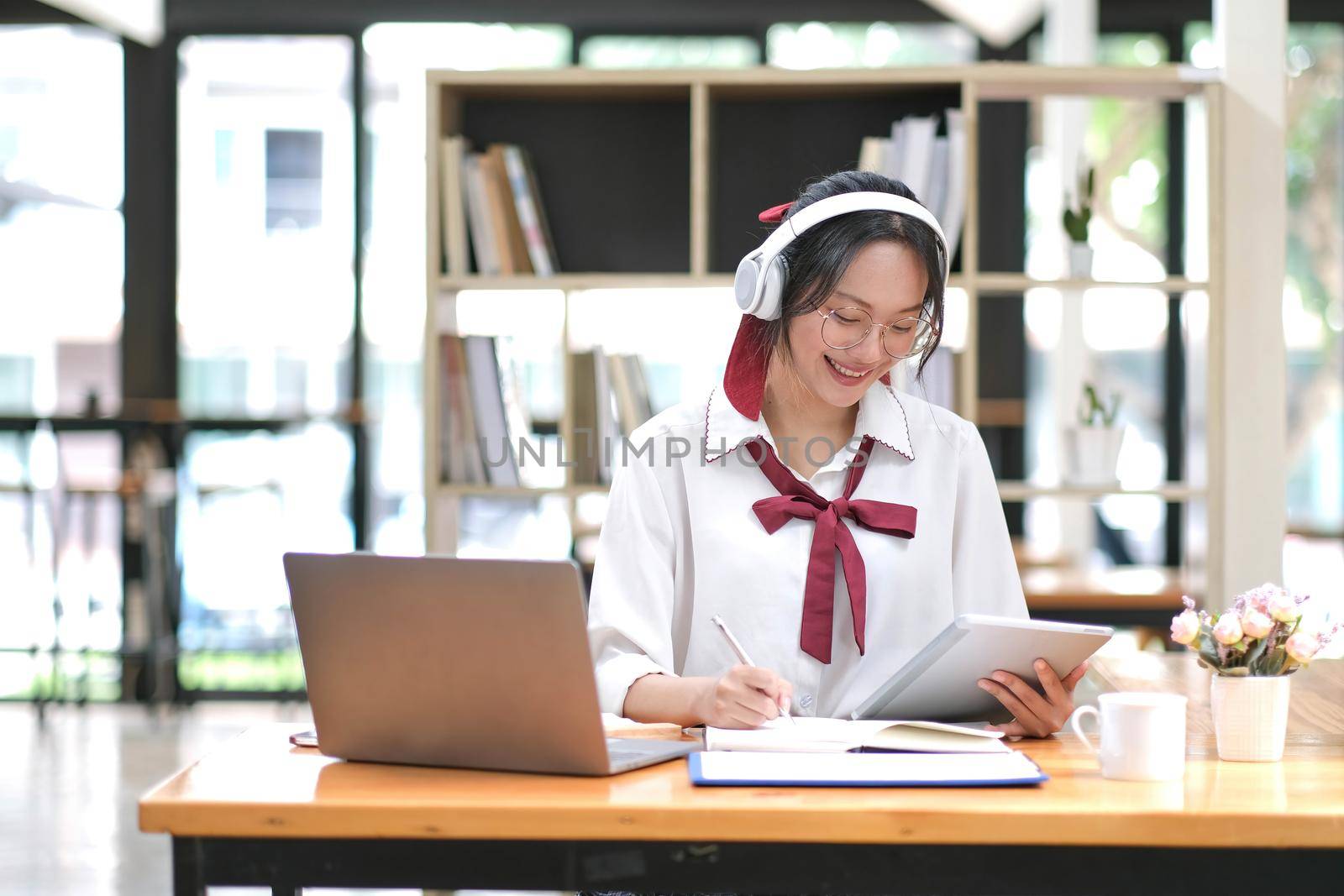 Happy young Asian student woman wearing headphones looking at webcam, looking at camera, during virtual meeting or video call talk. by wichayada