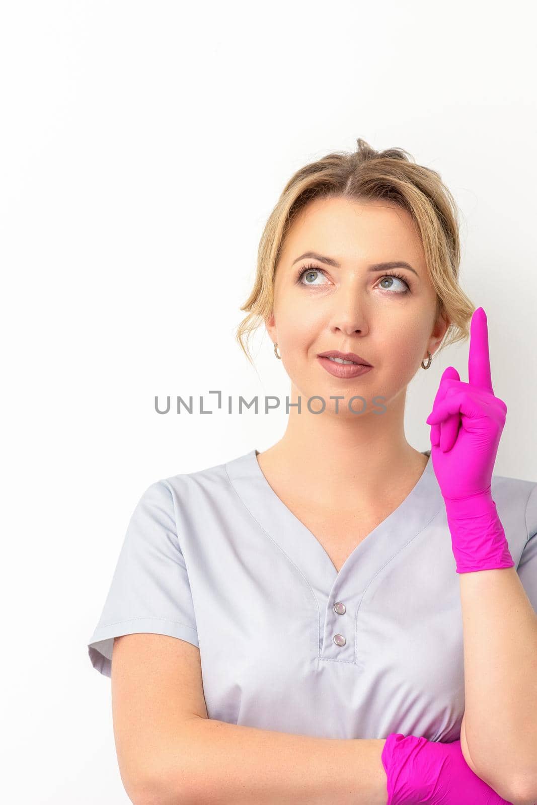 Portrait of young caucasian doctor woman wearing rubber gloves in medical uniform with one finger up, looking up against a white background, copy space. by okskukuruza