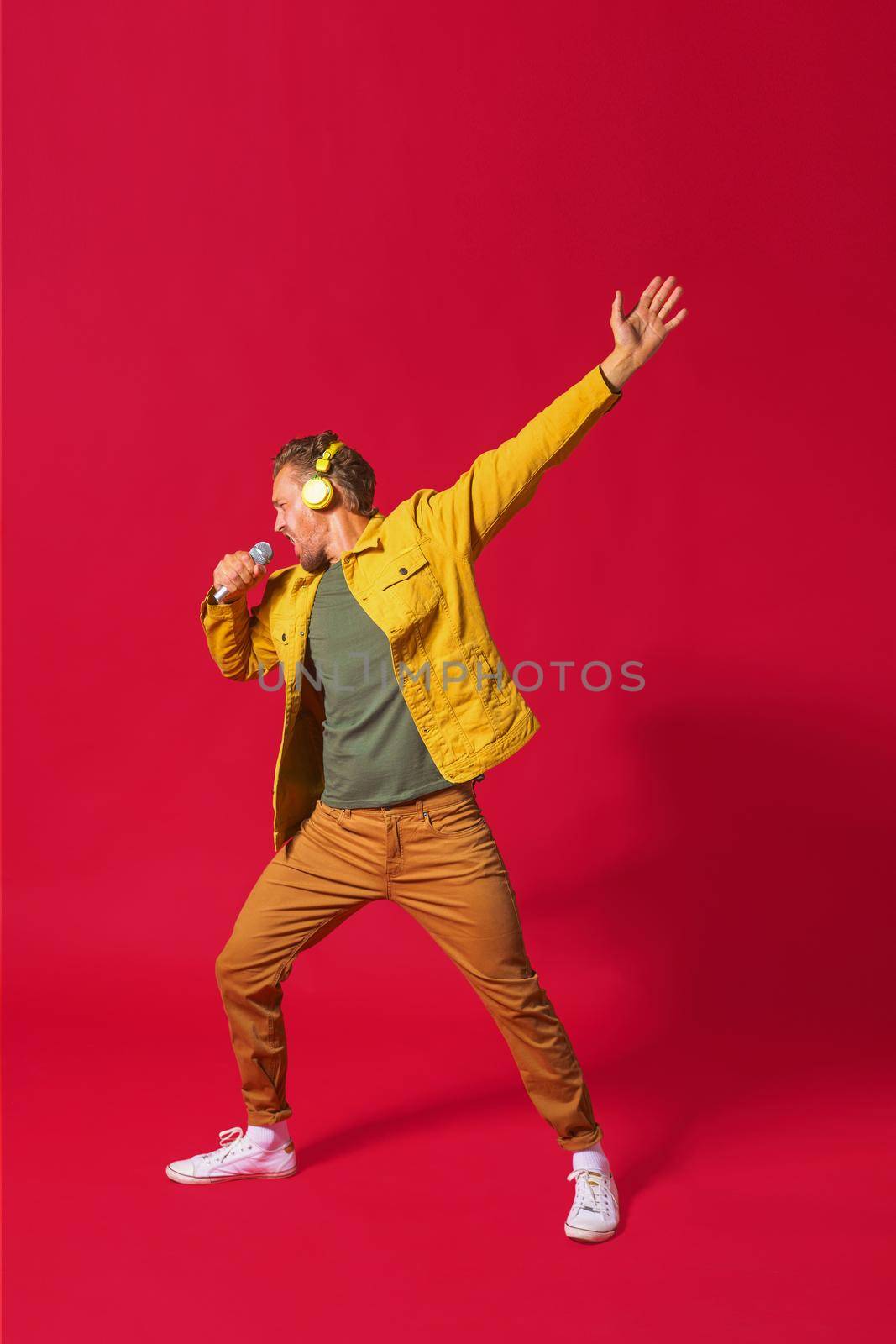 Singing young man enjoying song dancing with one hand up using phone and wireless headphones wearing jeans yellow jacket isolated on red background. Man sing while recording his voice by LipikStockMedia