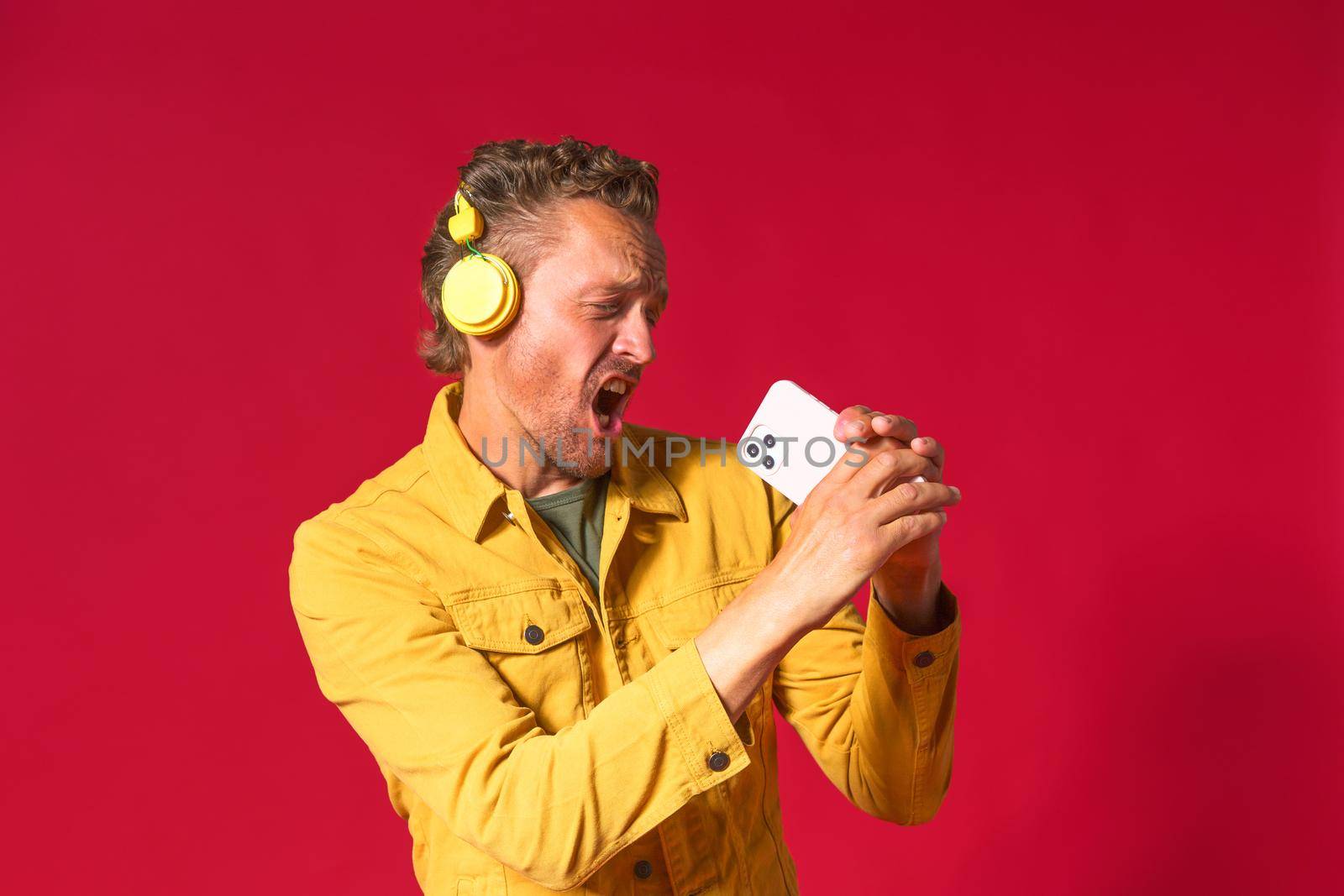 Singing handsome man enjoying his favorite song using phone and wireless headphones wearing jeans yellow jacket isolated on red background. Joyful man sing while recording his voice using smartphone.
