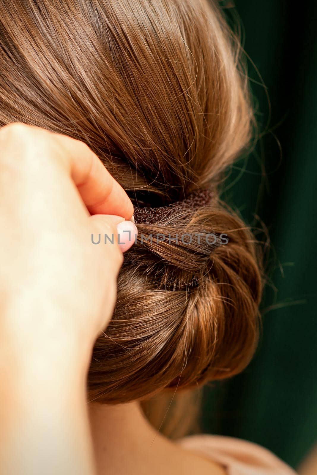 Hands of hairdresser making french twist hairstyle of an unrecognizable young brunette woman in a beauty salon, back view, close up. by okskukuruza