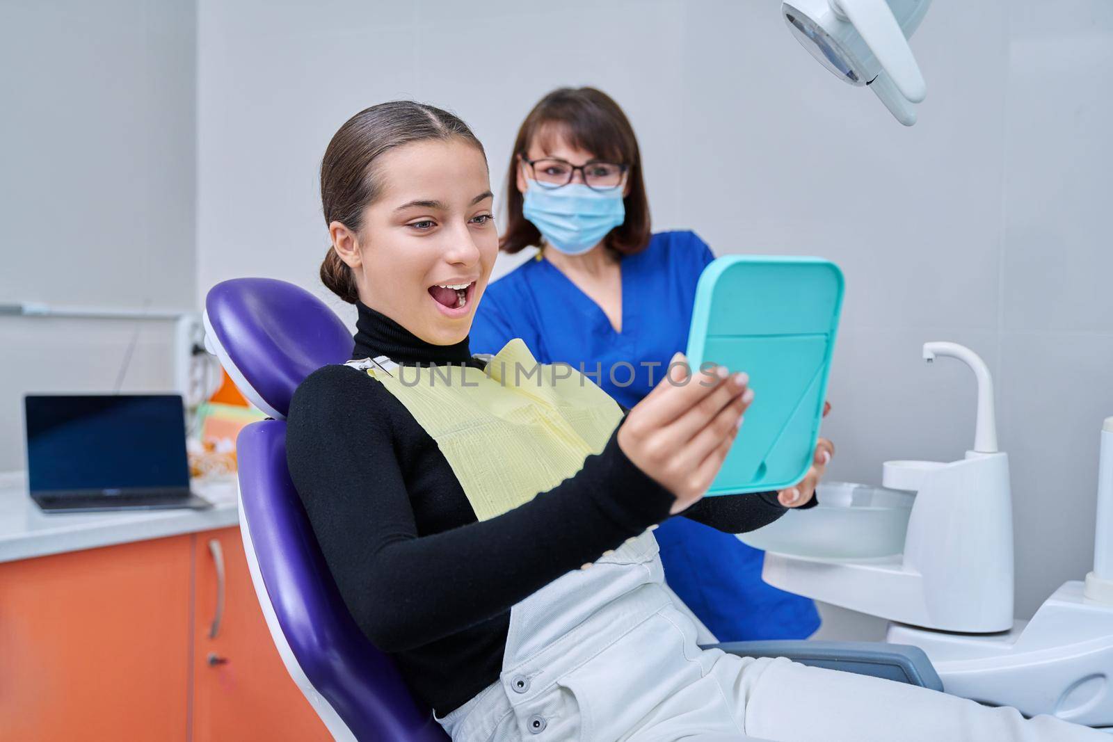 Happy young teenage female with mirror sitting in dentist chair smiling looking at healthy teeth, with dentist doctor in office. Dentistry, hygiene, treatment, dental health care concept