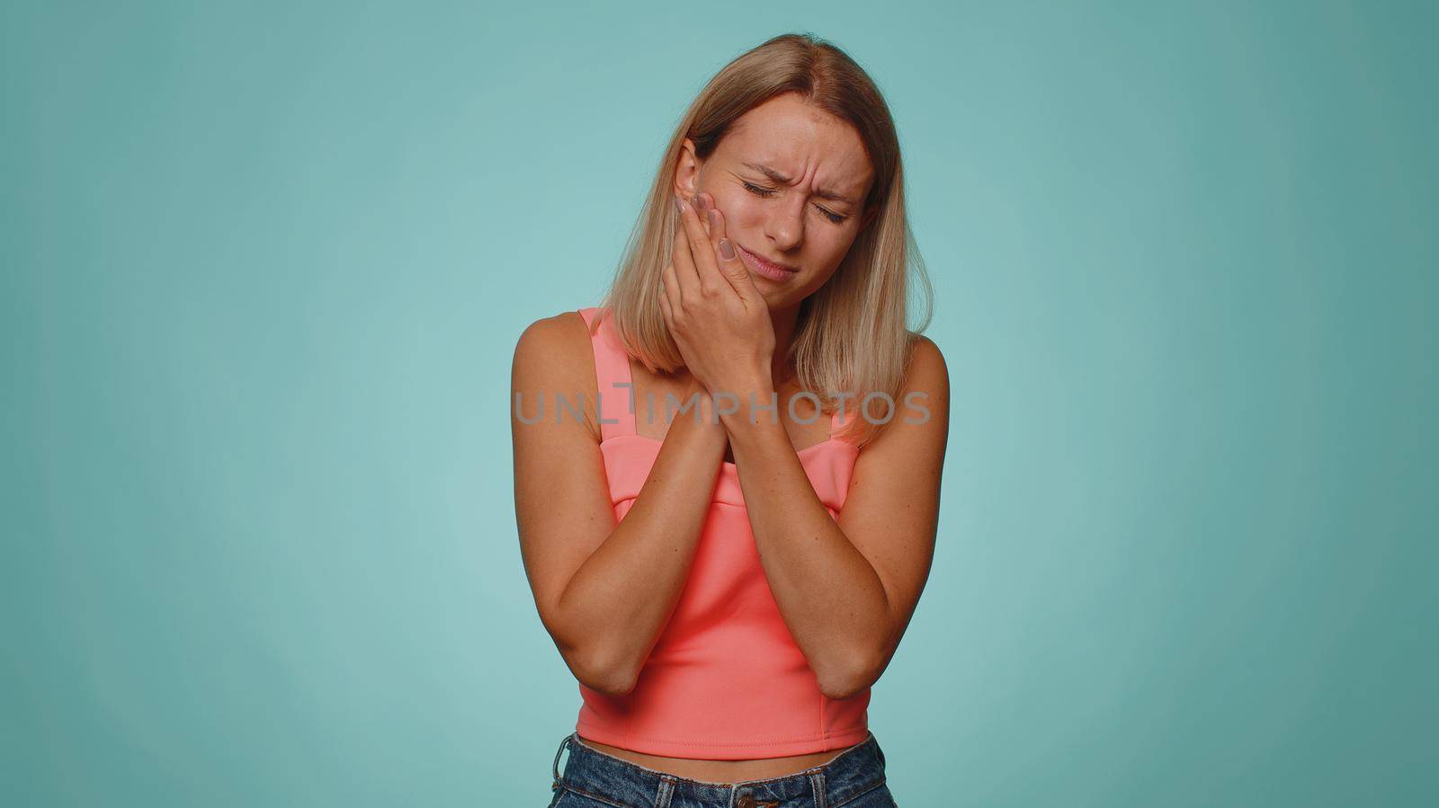 Blonde young woman touching sore cheek suffering from toothache cavities or gingivitis waiting for dentist appointment, gums disease. Adult girl indoors studio shot isolated alone on blue background