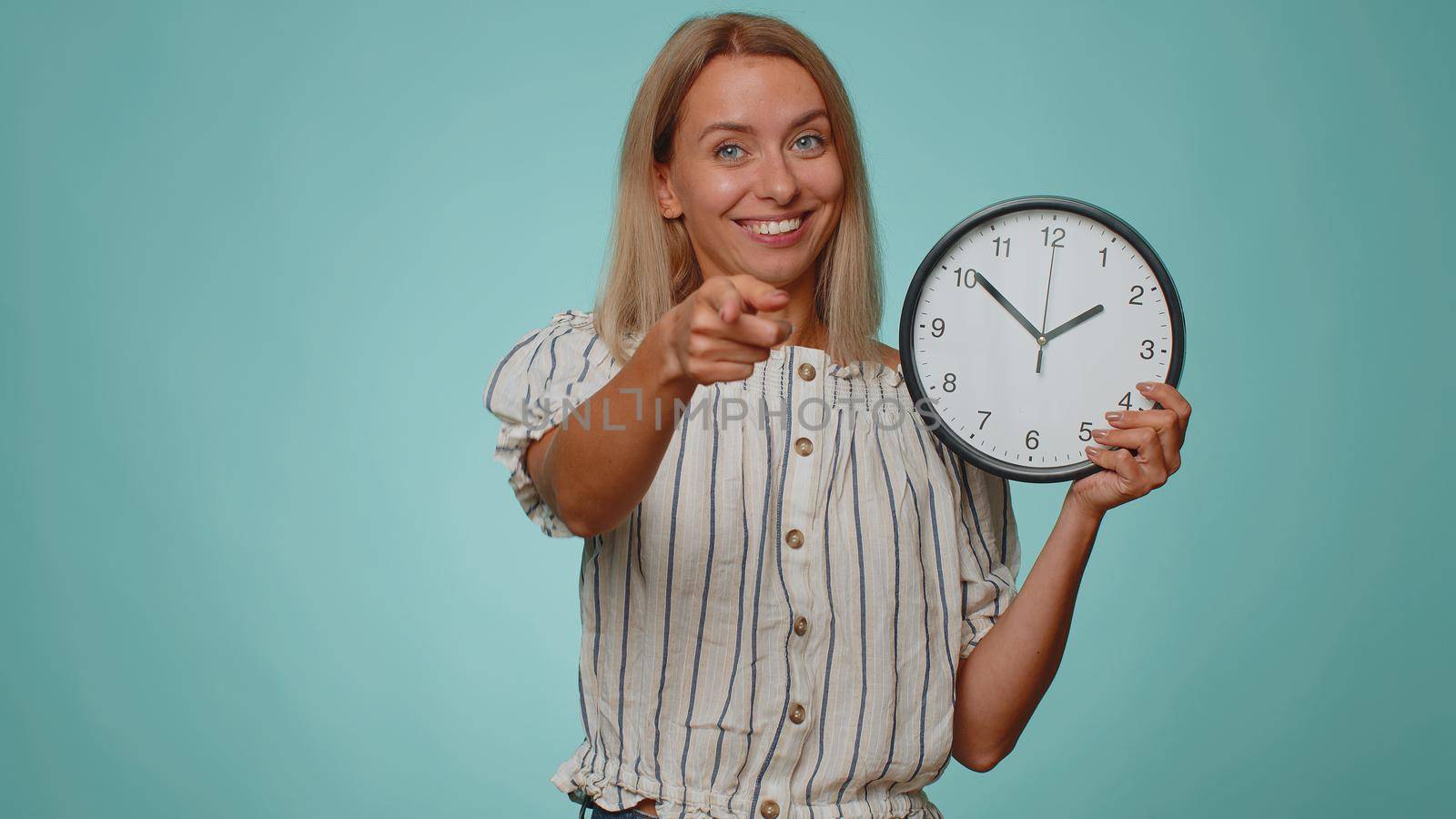 It is your time. Portrait of blonde young woman in shirt showing time on clock watch, ok, thumb up, approve, pointing finger at camera. Adult girl indoors studio shot isolated alone on blue background