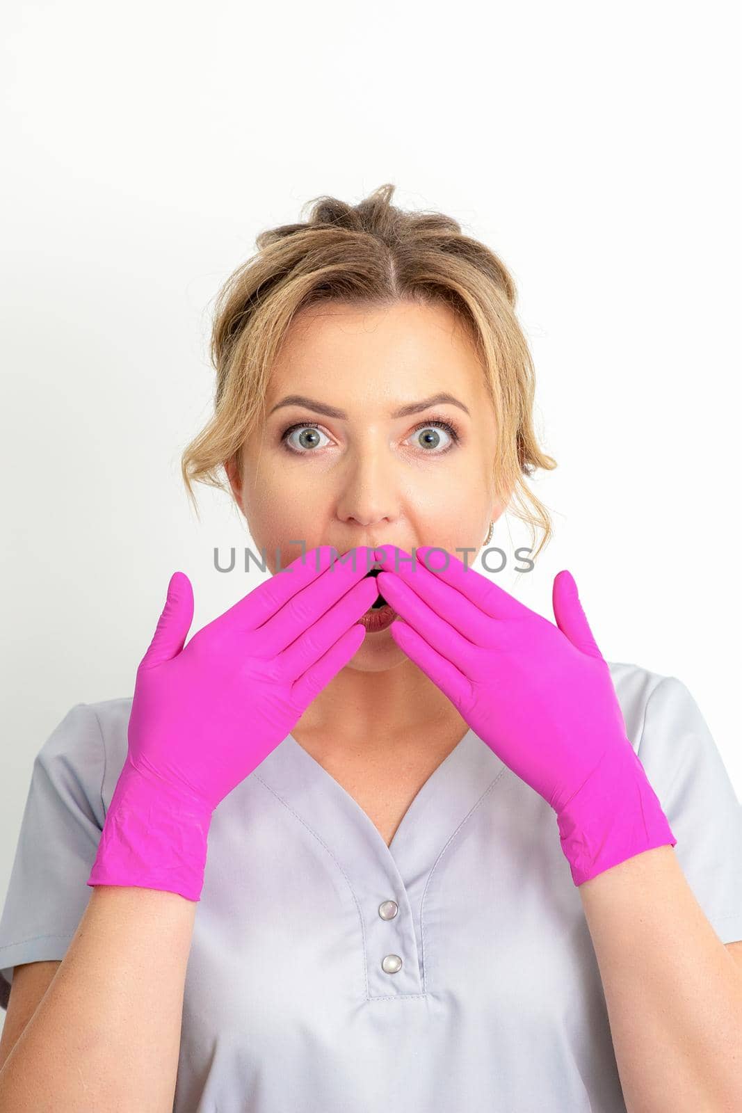 Portrait of a young female caucasian doctor or nurse is shocked covering her mouth with her pink gloved hands against a white background. by okskukuruza