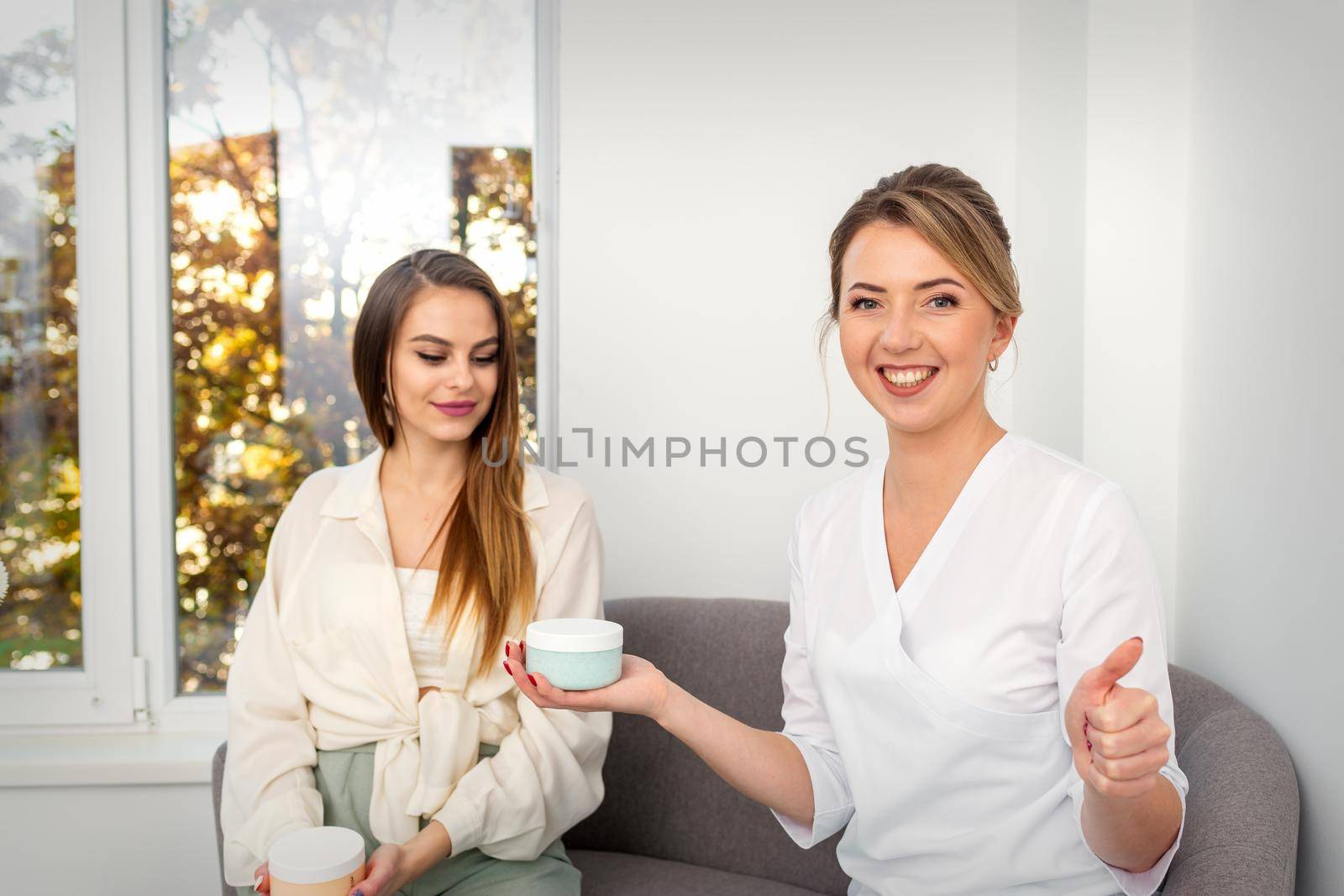 Beautician offering product for young woman, shows thumb up, holding a white plastic jar with a cream, mockup, copy space