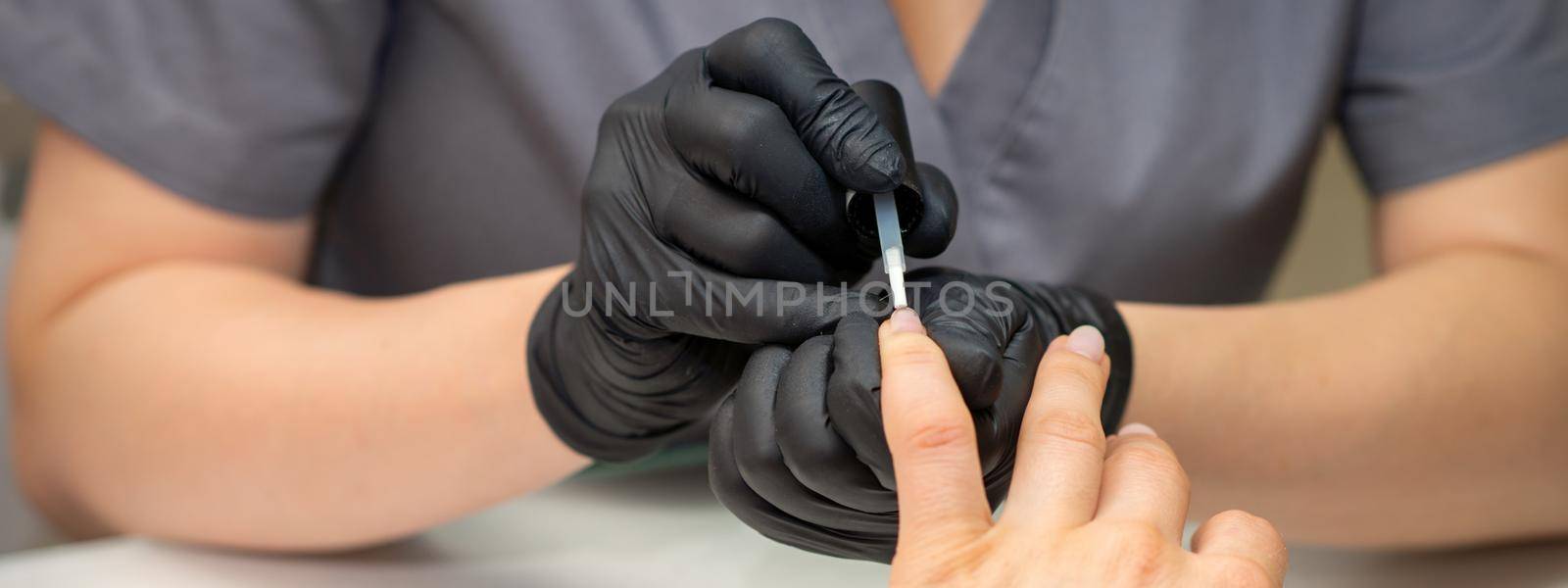 Painting female nails. Hands of manicurist in black gloves is applying transparent nail polish on female nails in a manicure salon. by okskukuruza
