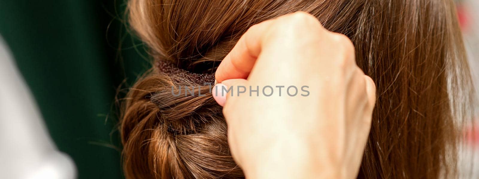 Hands of hairdresser making french twist hairstyle of an unrecognizable young brunette woman in a beauty salon, back view, close up