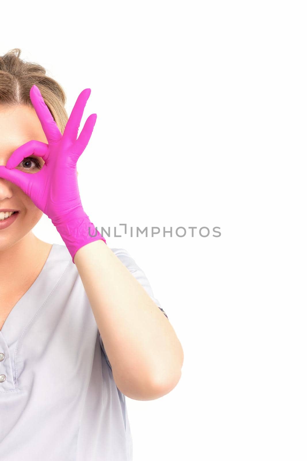 Smiling doctor oculist caucasian woman wearing pink rubber gloves in uniform showing ok sign covering the eye and thumb up gesture against a white background. by okskukuruza